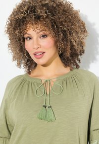 oliwkowa bluzka z okrągłym dekoltem, plisowanymi detalami i zapięciem na troczek z zielonymi chwostami. Gładka faktura, krótkie rękawy.