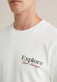 Balts kokvilnas T-krekls ar brīvu piegriezumu, uz kura ir uzraksts "Explore" drosmīgā pelēkā krāsā un "Your Horizon" sarkanā slīpā tekstā. Mīksta tekstūra, apaļā kakla dizains.