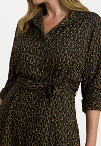 Rochie neagră cu model circular aurie, cu fesă butonat, guler și talie legată. Material moale, mâneci trei sferturi și un aspect finisat.