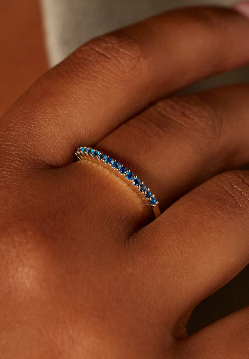Gold ring with a slim design, adorned with a row of small blue stones. The hand has a warm skin tone and textured details visible.
