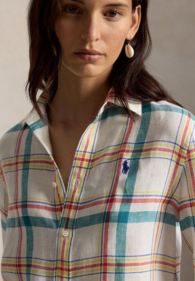 Kvinne iført en flerfarget rutete knappeskjorte med en blå brodert logo og en hvit skalløredobber på venstre øre.