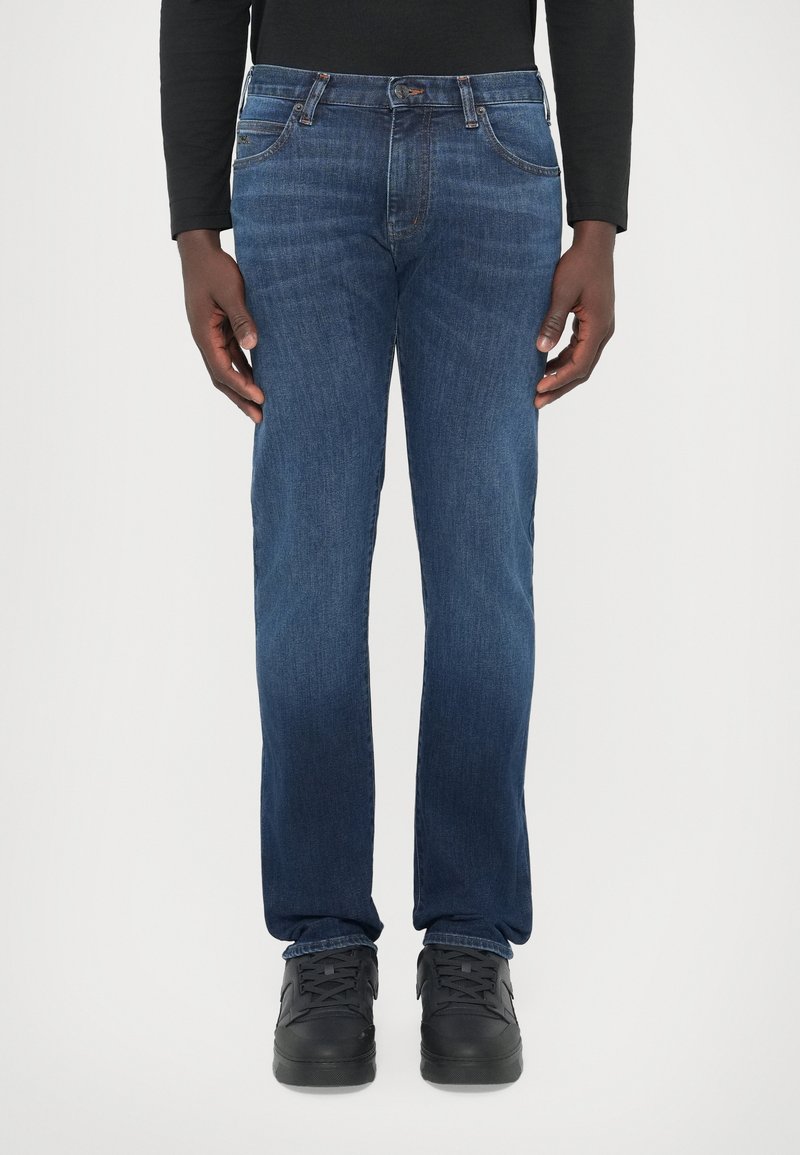 Vaqueros de mezclilla azul oscuro con corte recto, cinco bolsillos, sutil desgaste y costuras en contraste, combinados con zapatillas negras.