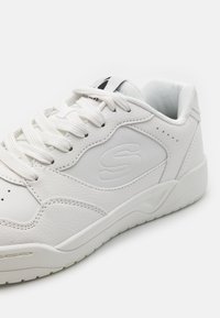 Zapatilla blanca de cuero con una textura suave, que presenta un logo lateral con patrón, cordones planos y una suela de goma. Acentos en gris sutil.