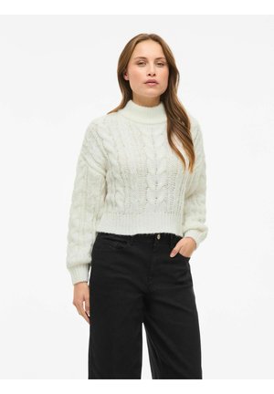 Bela pletena pulover s visokim ovratnikom in skrajšano zasnovo, v kombinaciji s črnimi hlačami visoke pasu. Vidna teksturirana tkanina.