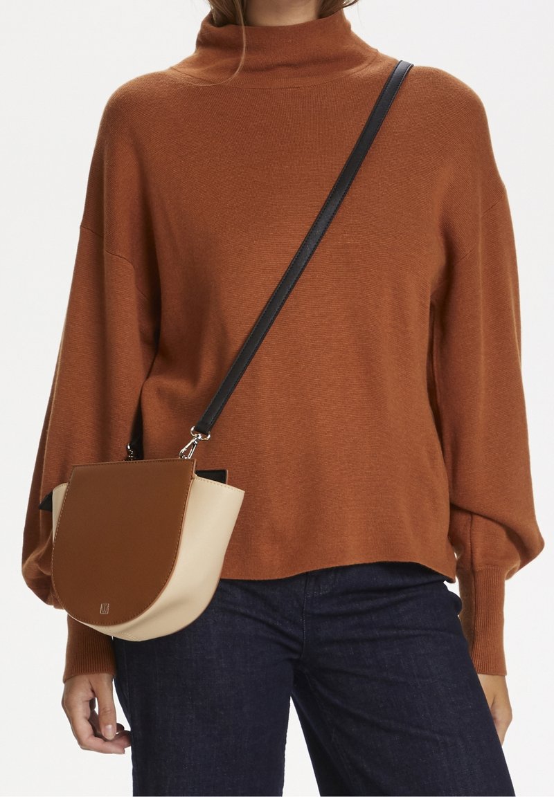 Femme portant un pull à col roulé marron et un jean foncé avec un sac bandoulière marron et beige avec une sangle noire.