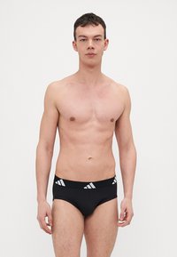 Schwarze Slips mit einem kontrastierenden weißen Gummibund, der drei diagonale Linien aufweist. Glatte Textur, enganliegendes Design, minimale Abdeckung.
