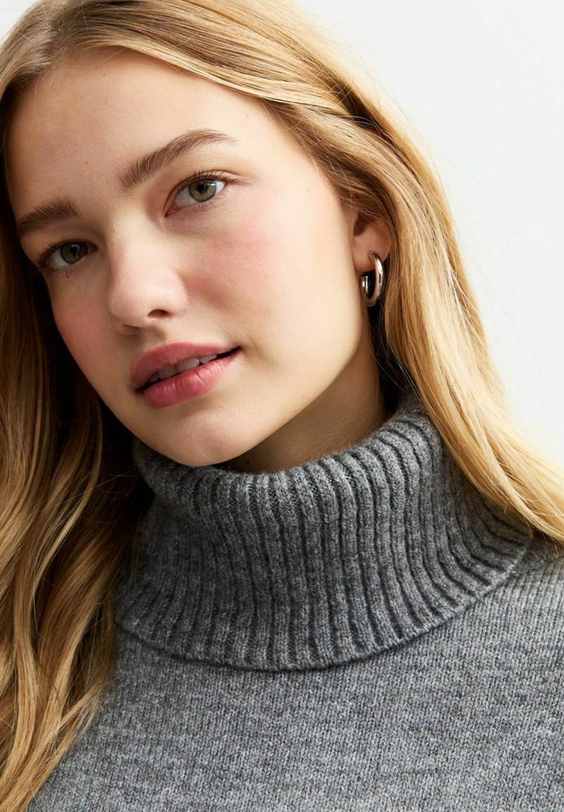 Grauer gerippter Rollkragenpullover aus weichem Strickstoff mit hohem Kragen und strukturierten Details. Das Modell hat lange, wellige Haare.