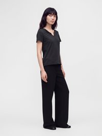 Woman with long dark hair wearing a black short-sleeve top and wide-leg black pants standing against a plain light background.