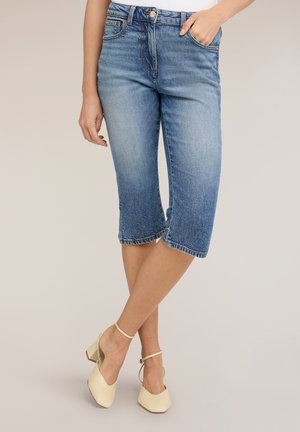 Blaue Denim-Knielange Shorts, getragen von einer Person mit einer Hand in der Tasche, gekreuzten Beinen und beigen Blockabsatz-Pumps mit Knöchelriemen.