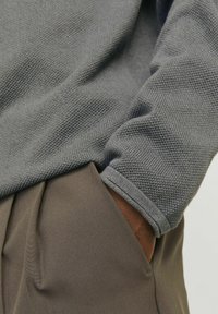 Gray textured sweater with a ribbed cuff, paired with brown pleated trousers, showing a hand in the pocket of the pants.
