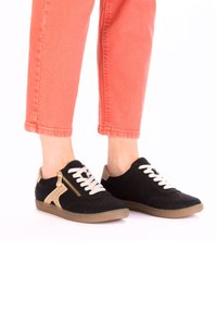 Black sneakers with a suede texture, cream accents, and a side zipper. Features white laces and a tan rubber sole, paired with coral pants.