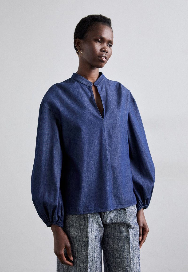 Navyblaues Denim-Oberteil mit Stehkragen, Knopföffnung und voluminösen Ärmeln. Getragen mit strukturierten grauen gemusterten Hosen.