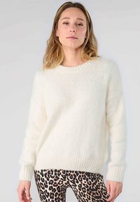 Weißer, flauschiger Pullover mit einem gerippten, runden Ausschnitt und langen Ärmeln, getragen über beige-schwarzen Leggings mit Leopardenmuster.