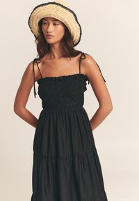 Abito nero a più livelli con corpetto arricciato e spalline sottili. Abbinato a un cappello di paglia a tesa larga con dettaglio nero.