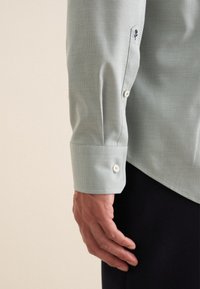 Camicia testurizzata verde chiaro con colletto a punta, polsini con bottone e un accento ricamato sulla manica. Presenta chiusure a bottone bianche.
