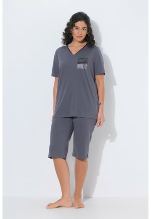 Frau mit lockigem dunklem Haar, die ein lockeres graues V-Ausschnitt-Oberteil mit einer glänzenden Brusttasche und passenden knielangen Shorts trägt, barfuß auf weißem Boden.