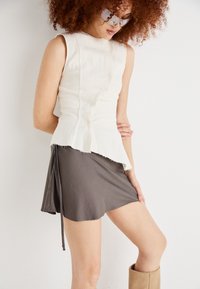 White sleeveless top with a pleated texture and frayed hem; paired with a gray mini skirt featuring a tie detail. Light brown knee-high boots.