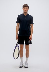 Μαύρο πόλο πουκάμισο με κοντά μανίκια, κεντημένο λογότυπο, σετ με μαύρο σορτς και παπούτσια τένις, κρατώντας ρακέτα τένις με μοβ λεπτομέρειες.