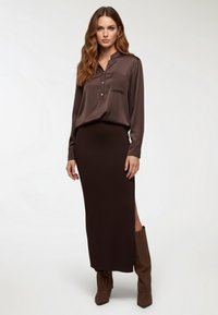Blusa lunga manica in satin marrone con colletto e taschino, abbinata a una gonna marrone aderente con spacco laterale, indossata con stivali marroni al ginocchio.