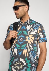 Πουκάμισο με κοντά μανίκια και κουμπιά, με πολύχρωμο αφηρημένο λουλουδάτο σχέδιο σε σκούρο φόντο· διαθέτει γιακά και τσέπη στο μπροστινό μέρος.