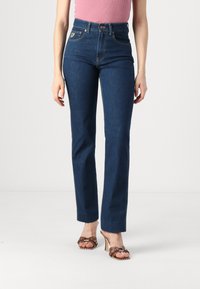 Dark blue high-waisted jeans feature a straight leg design, five pockets, gold stitching, and a branded patch on the back.