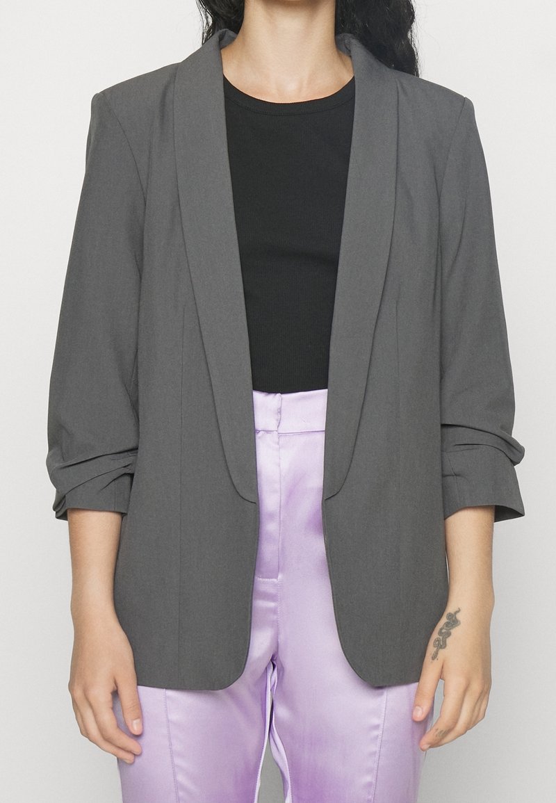 Personne portant un blazer gris avec les manches retroussées, un top noir et un pantalon violet clair brillant, montrant un petit tatouage sur le poignet de la main droite.