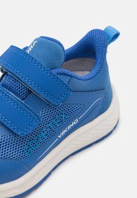 Modrá teniska s priedušným a syntetickým zvrškom, vybavená suchým zipsom, textúrovaným dizajnom a bielou gumovou podrážkou s embosovaným logom.