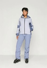 Γυναίκα με ανοιχτό μπλε και γκρι μπουφάν σκι, ταιριαστά παντελόνια σκι, μαύρα μποτάκια σκι και μαύρα γυαλιά σκι στο κεφάλι της, στέκεται σε εσωτερικό χώρο.