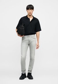 Νεαρός άνδρας όρθιος, φοράει μαύρο πόλο με κοντά μανίκια, ανοιχτό γκρι στενά τζιν, μαύρα αθλητικά παπούτσια, κρατώντας μια μαύρη τσάντα κάτω από το μπράτσο του.