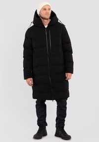Giacca a vento nera con silhouette lunga, chiusura con zip, cappuccio regolabile e due tasche frontali, indossata sopra jeans blu e stivali neri.