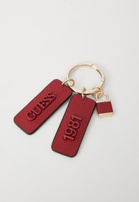 Rød struktureret nøglering med to mærker: et med "GUESS" præget og det andet med "1981." Fastgjort med ring i guldtonet og en lille lås charm.