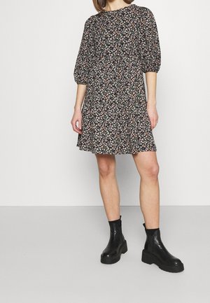 Kvinde iført en sort knælængde kjole med blomsterprint med pufærmer til albuen og sorte chunky ankelstøvler.