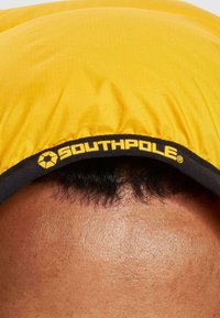 Geltonas audinio galvos apdangalas su juodu apvadu, kuriame yra geltona "SOUTHPLOLE" logotipo ženklo. Tekstūra atrodo lygi su nedideliu blizgesiu.