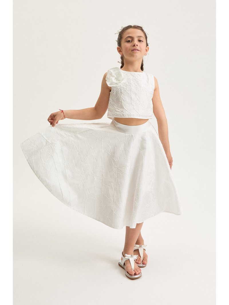Ung pige i en hvid tekstureret ærmeløs top med blomstdetalje og matchende vid nederdel, poserende mens hun holder nederdelen ud.