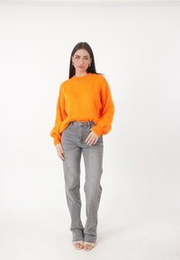 Eine Frau steht posierend da, trägt einen leuchtend orangefarbenen Pullover, graue Jeans, weiße Sandalen mit offenen Zehen und goldene Creolen vor einem weißen Hintergrund.