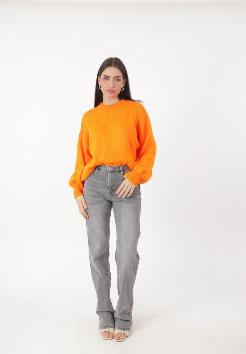 Donna in piedi in posa, che indossa un brillante maglione arancione, jeans grigi, scarpe con tacco aperte bianche e orecchini a cerchio dorati su uno sfondo bianco.
