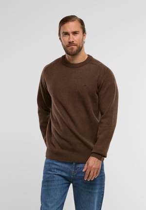 Brauner Strickpullover mit Rundhalsausschnitt, gerippten Bündchen und Saum; verfügt über ein kleines Logodetail auf der Brust, getragen über blauen Jeans.