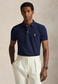Mann trägt ein marineblaues kurzärmeliges Poloshirt mit weißem Logo und cremefarbenen, hoch taillierten Faltenhosen und steht mit einer Hand in der Tasche.