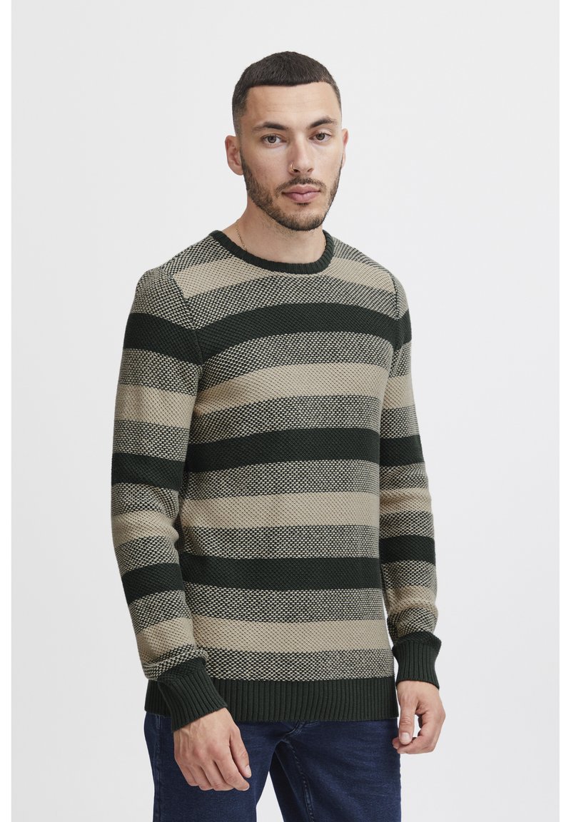 Zeleni in bež svilen pulover z teksturirano pletenino. Ima okroglo ovratnik in rebraste manšete ter rob. Obut je z temno modrimi jeans hlačami.