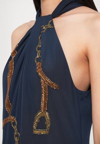 Rochie tip halter bleumarin cu broderie elaborată din mărgele bronzate, cu modele de lanțuri și scărițe, evidențiind textura netedă a țesăturii și căderea acesteia.