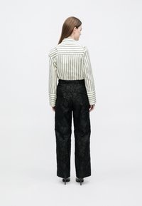 Mulher em pé, virada de costas, vestindo uma camisa branca às riscas e calças pretas texturizadas de cintura alta, com sapatos pretos de salto.