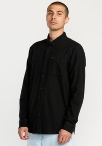 Chemise noire à boutons en tissu doux, avec une poche poitrine, un col standard et des manches longues. Branding minimal sur la poche.