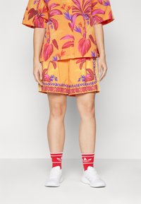 Lys oransje blomster skjorte med rosa og blå mønstre, kombinert med matchende oransje shorts med dekorativ kant. Komplementert av hvite sneakers og røde sokker.