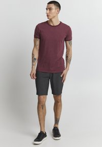 Maroon t-shirt with short sleeves and a round neck, paired with dark gray shorts. Worn with black sneakers, featuring visible tattoos.