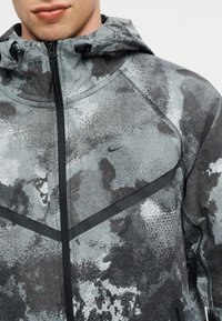 Κοντινό πλάνο ενός ατόμου που φορά γκρι και μαύρο μπουφάν με κουκούλα σε καμουφλάζ σχέδιο, μαύρο φερμουάρ και μικρό λογότυπο Nike στο στήθος.