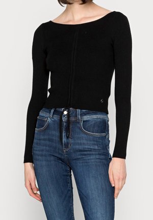 Kvinde iført en tætsiddende sort ribstrikket langærmet top med en let udskæring og blå højtaljede jeans mod en ensfarvet baggrund.