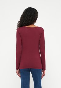 Langærmet bordeaux top i et blødt, glat stof med rund halsudskæring og tætsiddende pasform. Sammen med blå jeans.