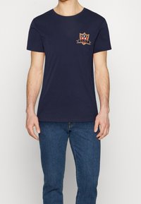 Camiseta de algodón azul marino con cuello redondo, que presenta un logo de escudo bordado en rojo, blanco y dorado en el lado izquierdo del pecho. Ajuste clásico.