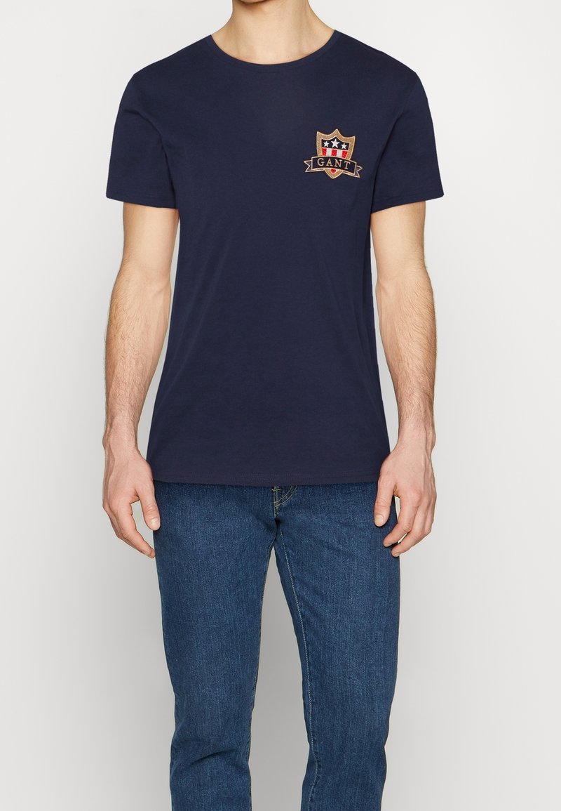 Camiseta de algodón azul marino con cuello redondo, que presenta un logo de escudo bordado en rojo, blanco y dorado en el lado izquierdo del pecho. Ajuste clásico.