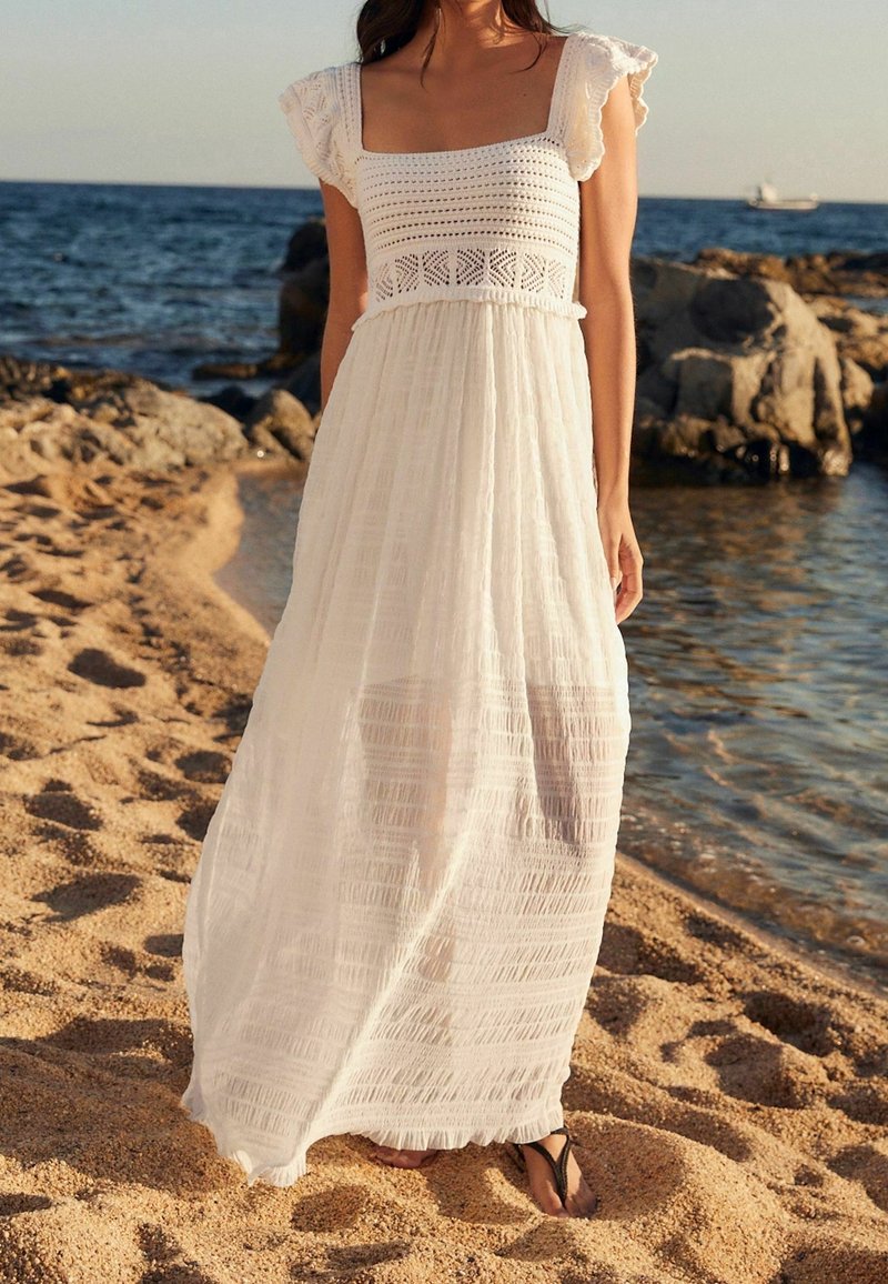 Femme debout sur une plage de sable près des rochers et de la mer, portant une longue robe blanche avec un haut en crochet et des manches volantées.